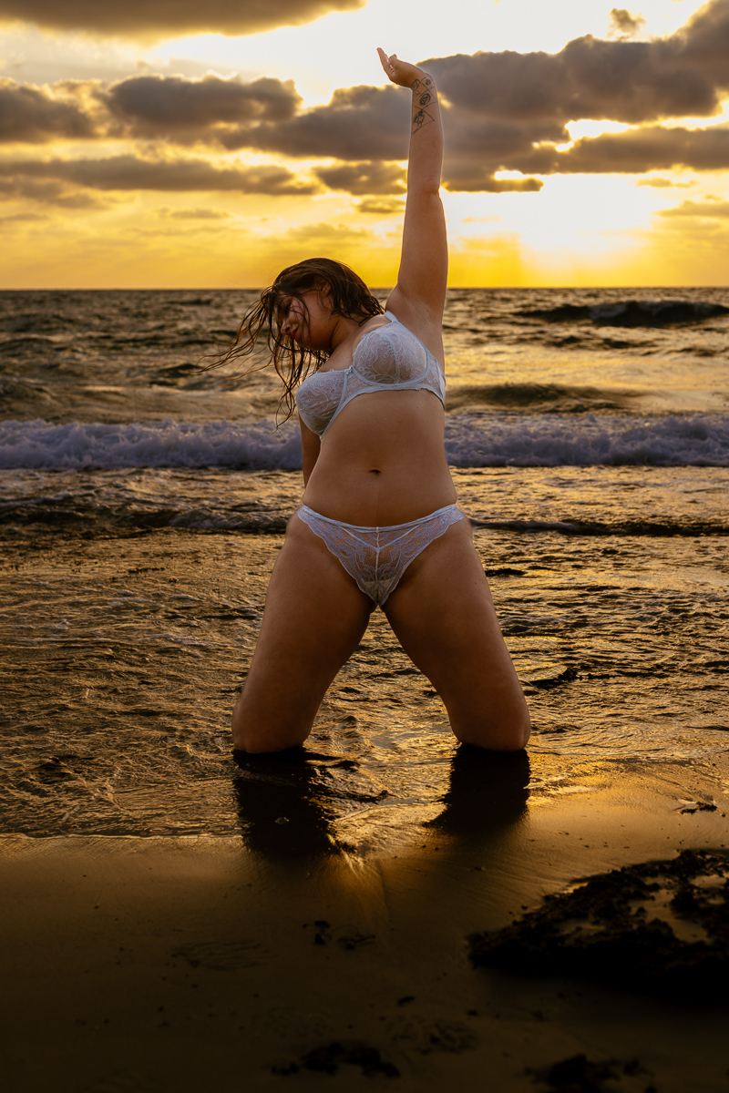 Gorgeous woman in a powder-blue bra and panties set on the beach, stretching way back towards the sunset, left arm thrust to the sky.