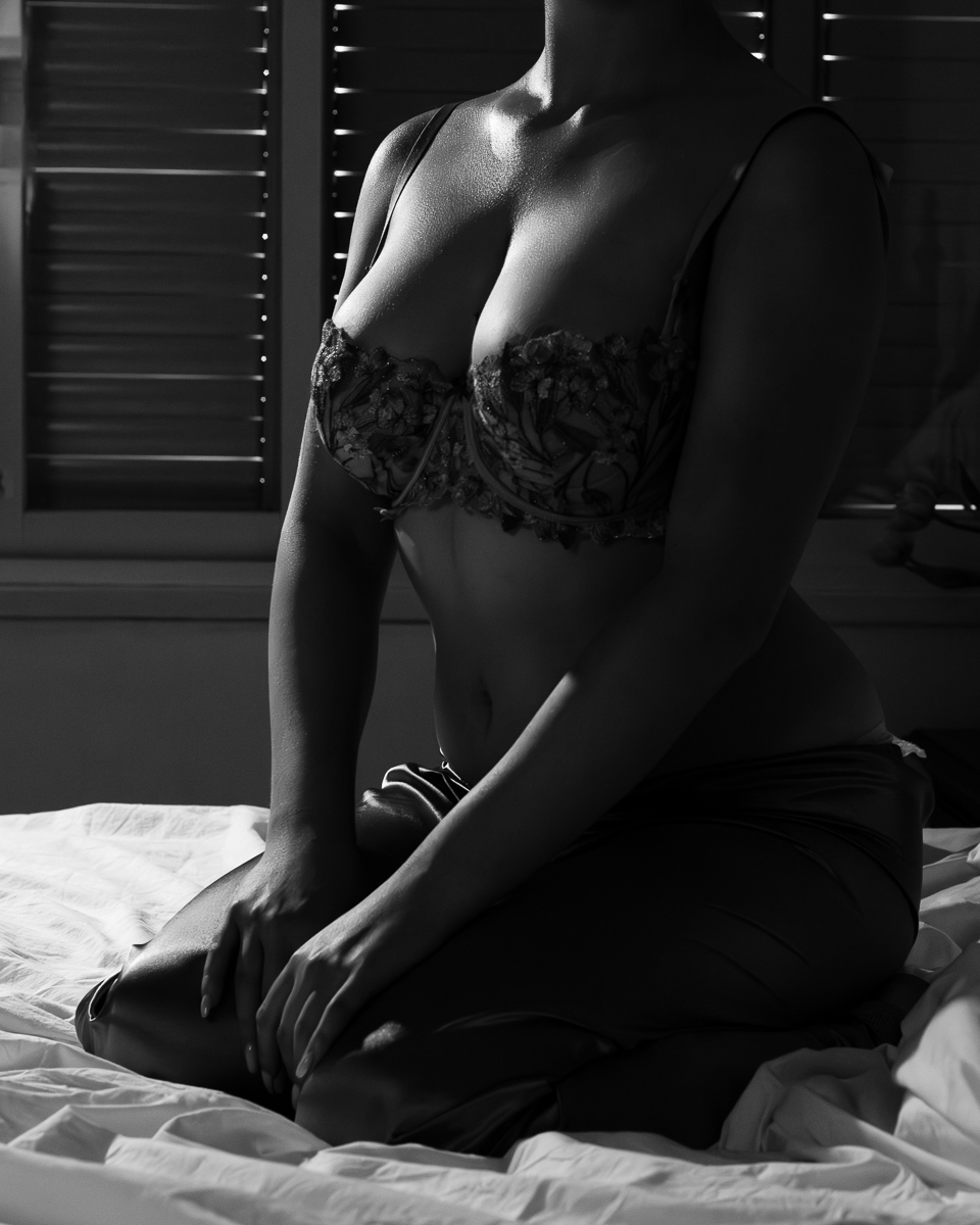 Black and white image of a woman, from her neck down, kneeling on a bed, light playing beautifully across her breasts, her neck, and her forearms and thighs.
